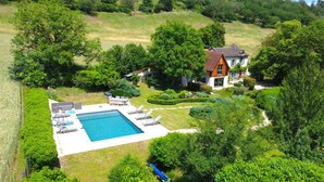 Una amplia piscina con tumbonas, rodeada de jardines cuidados y una casa con techo inclinado.