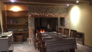 Un comedor con chimenea de piedra, sillas de madera, una mesa preparada para cuatro personas y una estantería con libros.