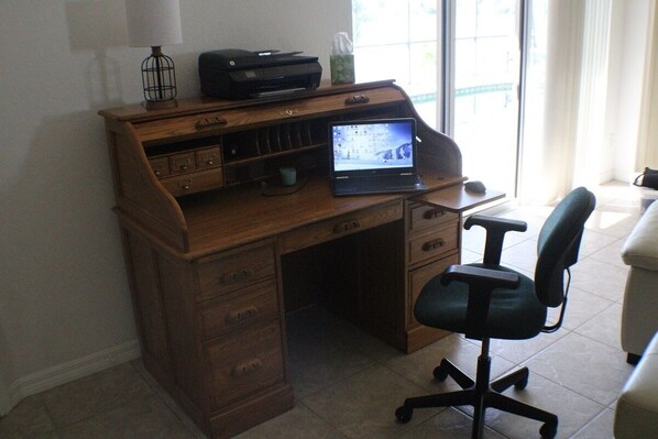 Un escritorio de madera con una computadora portátil, una impresora y una lámpara. Hay una silla y un ventanal con cortinas.