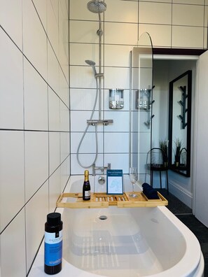 A modern bathroom with a freestanding bathtub, a showerhead, and a vanity with a mirror. There is a bottle of shampoo and a glass of water on the vanity.