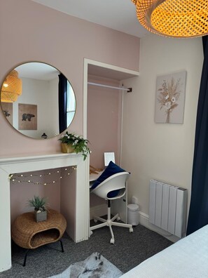 A bedroom with a bed, a desk, a chair, a radiator, a mirror, and a wall-mounted shelf with a plant.