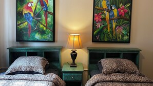 Una habitación con dos camas, cada una con una colcha de estilo leopardo, flanqueadas por mesitas de noche verdes. En la pared, dos cuadros enmarcados con loros de colores.