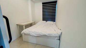 A small, neatly arranged bedroom with a single bed, a bedside table, and a window with blinds.