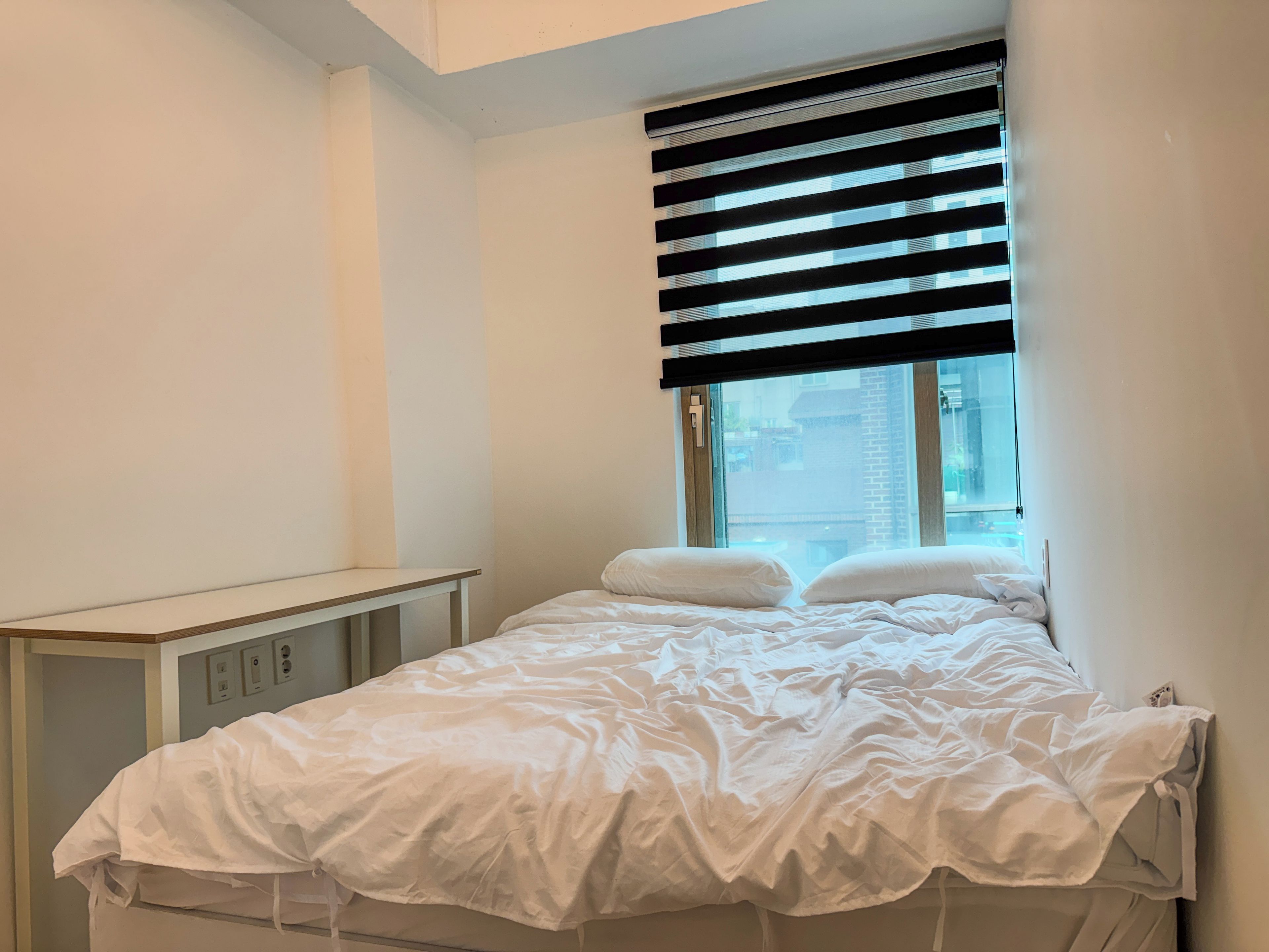 A bedroom with a bed, a window with blinds, a bedside table, and a wall-mounted control panel.