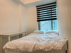 Ein Schlafzimmer mit einem Bett, einem Fenster mit Jalousien, einem Nachttisch und einer Wandbedienung.