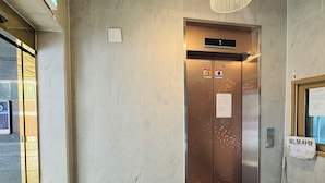 Un ascensor con pantalla digital, señal de salida de incendios y un cartel con texto en coreano.