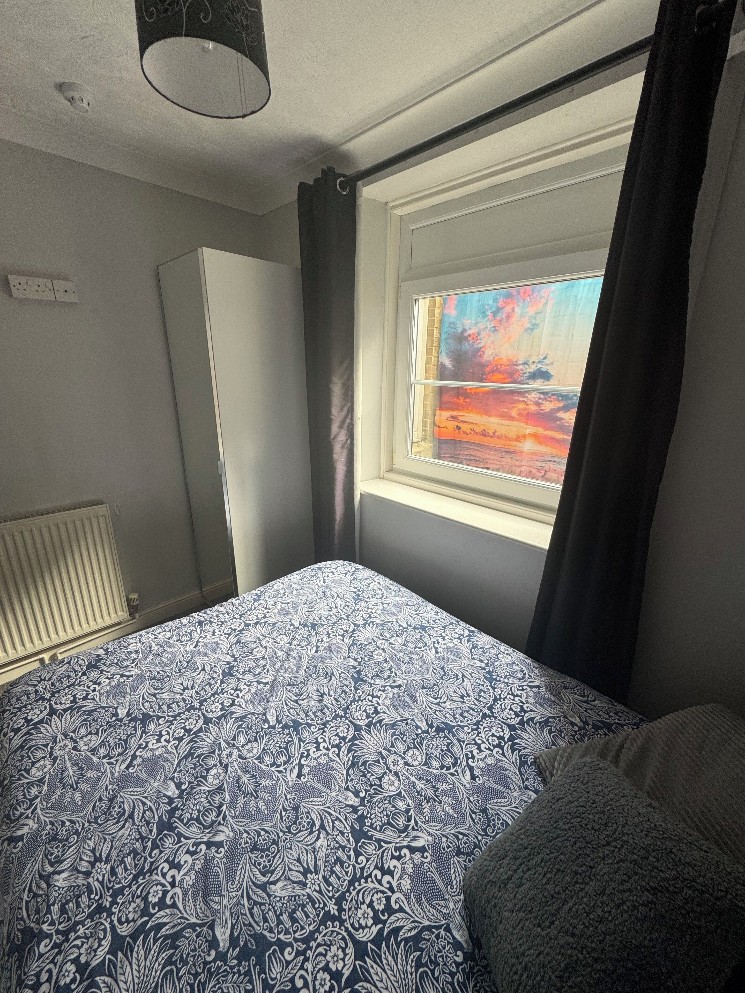 Une chambre à coucher avec un lit, une fenêtre donnant sur un coucher de soleil, un radiateur et une armoire.