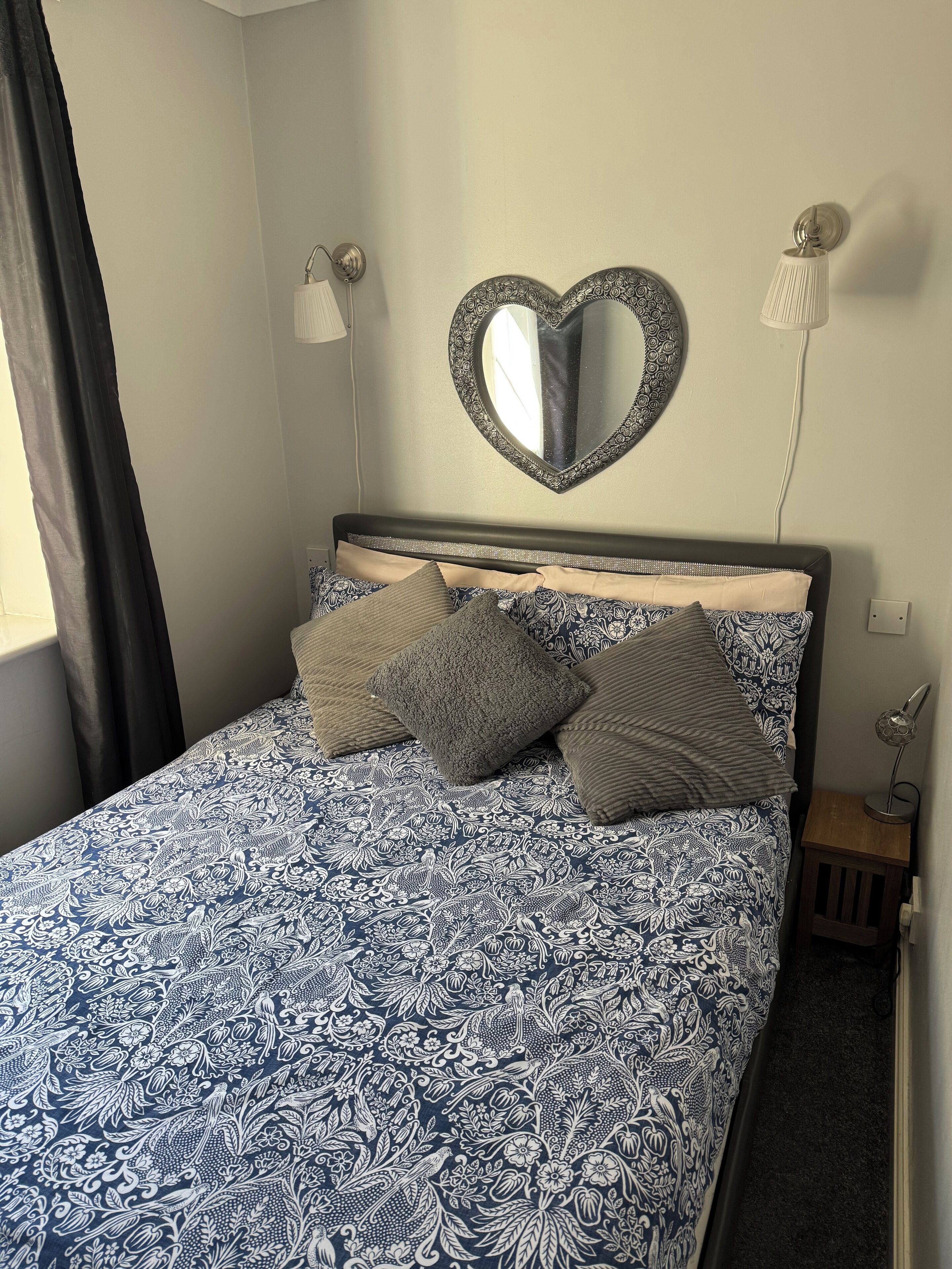 A bed with a patterned bedspread, a heart-shaped mirror above it, and two bedside lamps.