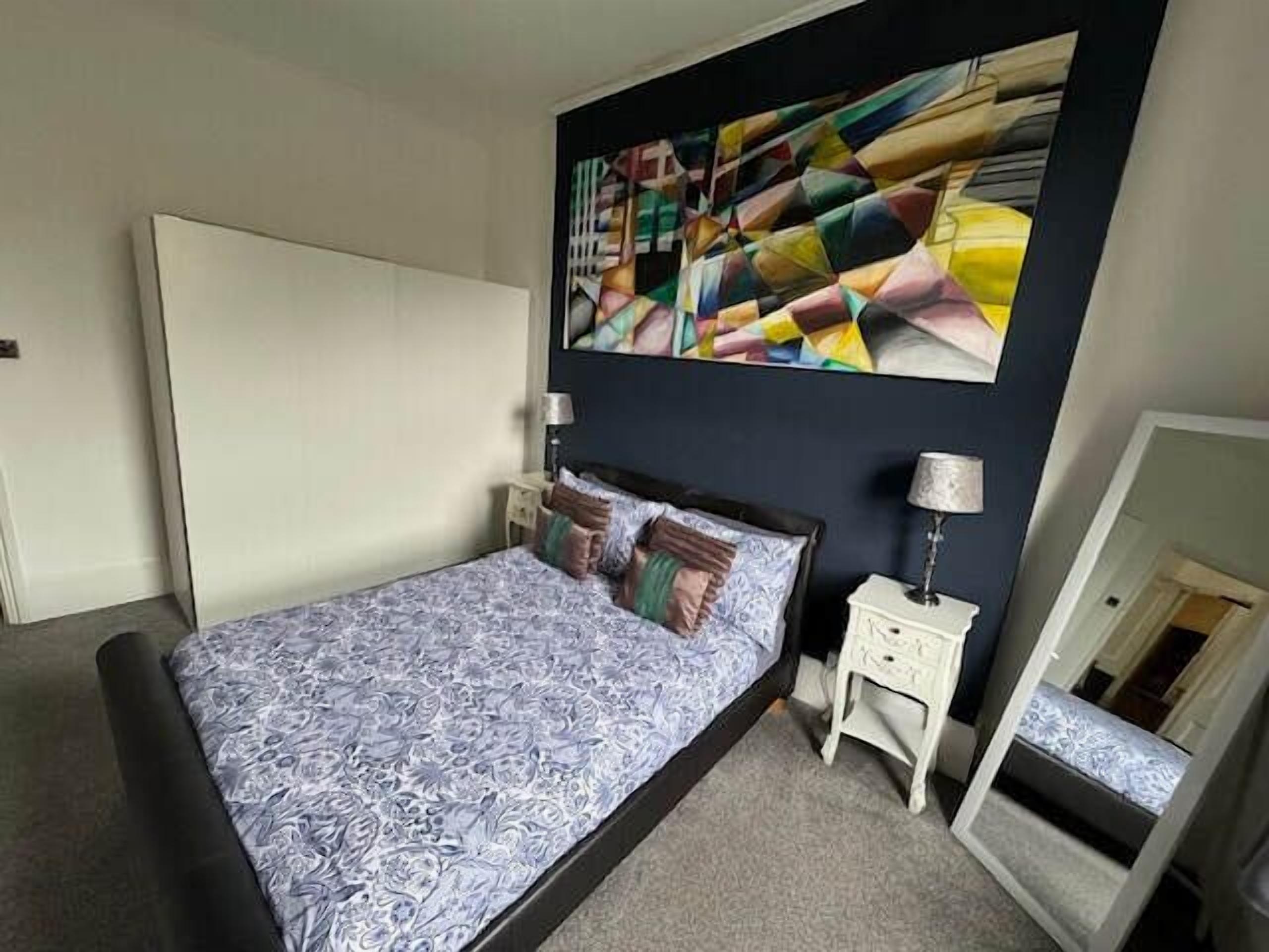 Een slaapkamer met een bed, een nachtkastje, een lamp, een spiegel en een groot abstract schilderij aan de muur.