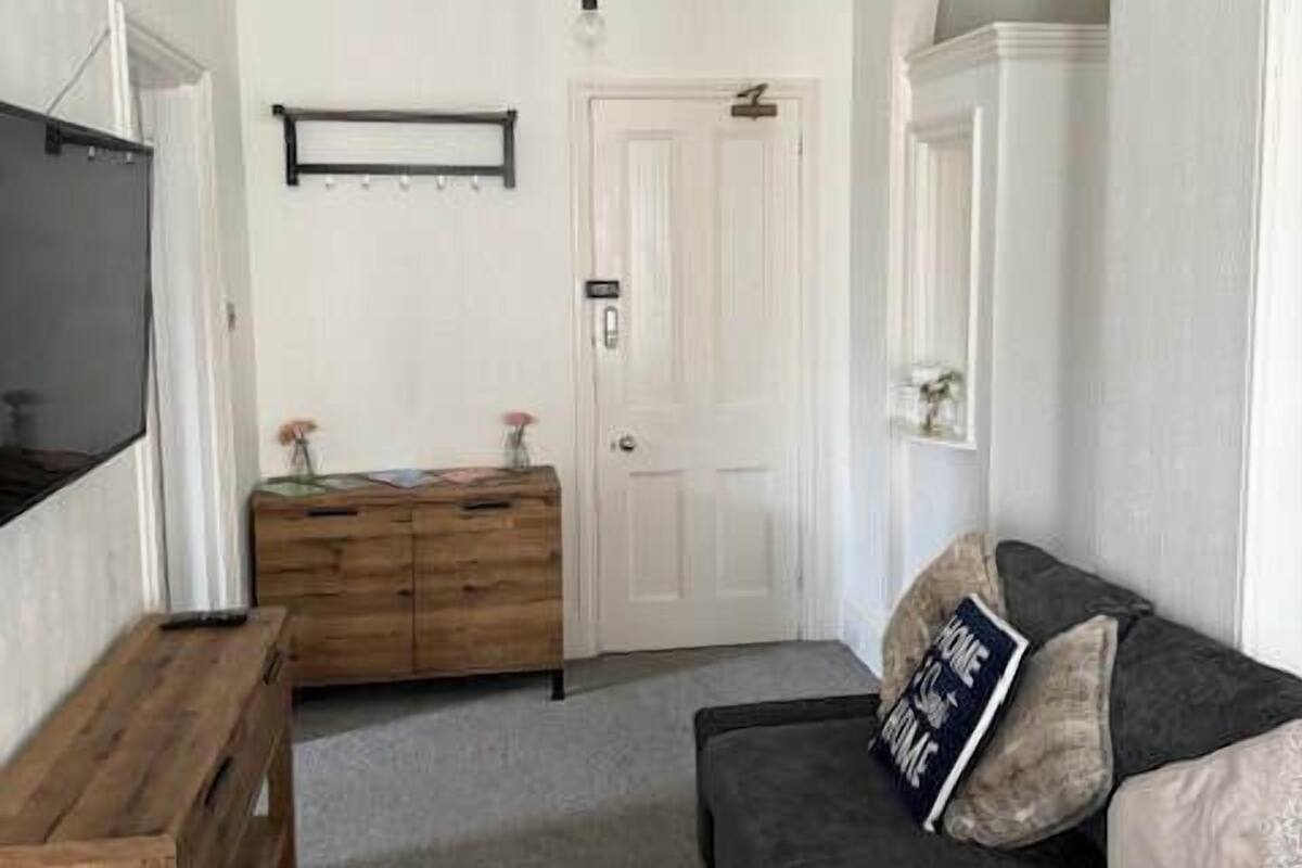 Un salon avec un canapé foncé, une table d’appoint en bois et un téléviseur à écran plat fixé au mur.