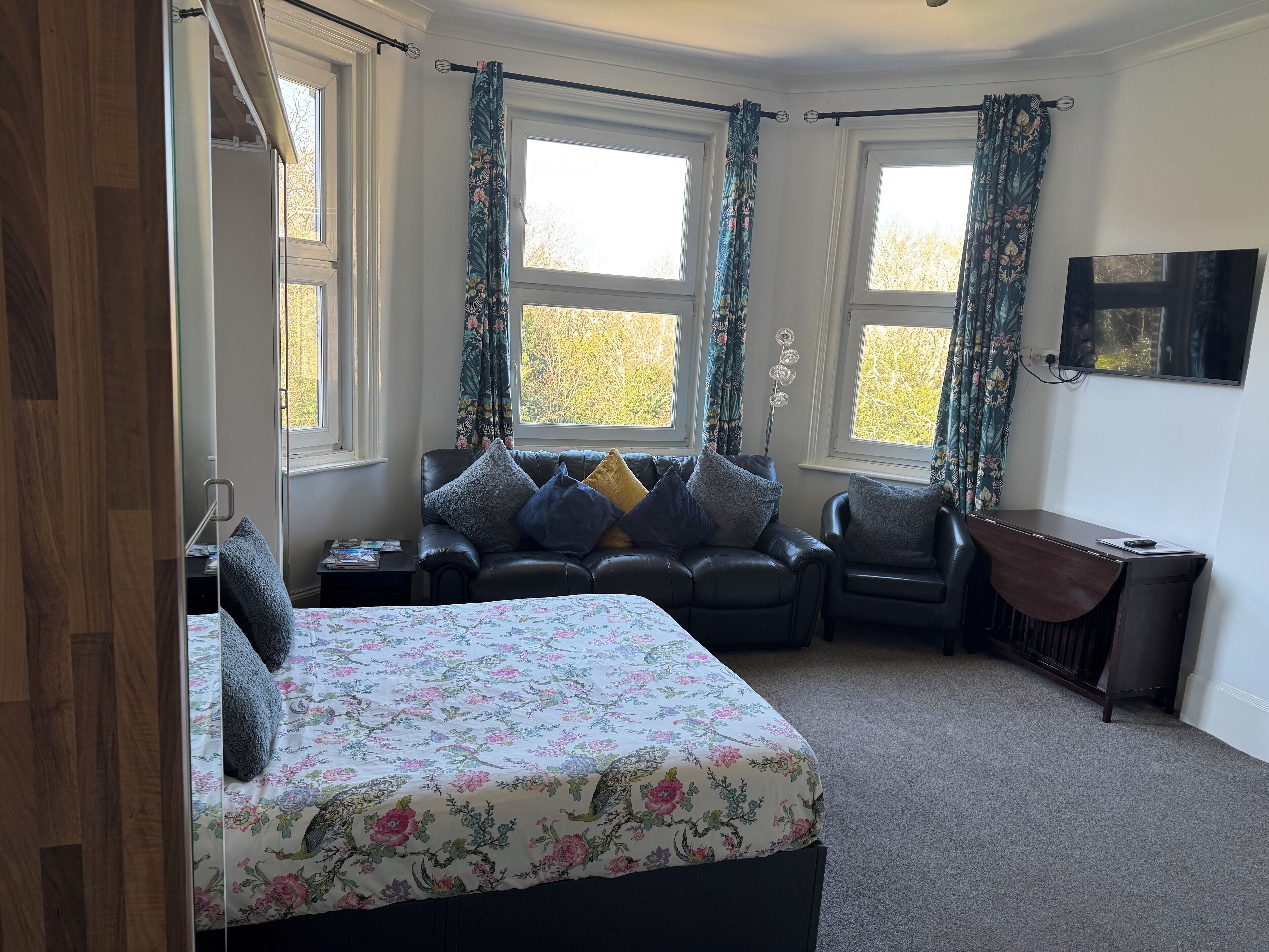 A bedroom with a bed, a desk, and two chairs. There are windows with curtains, a mirror, and a wall-mounted TV.