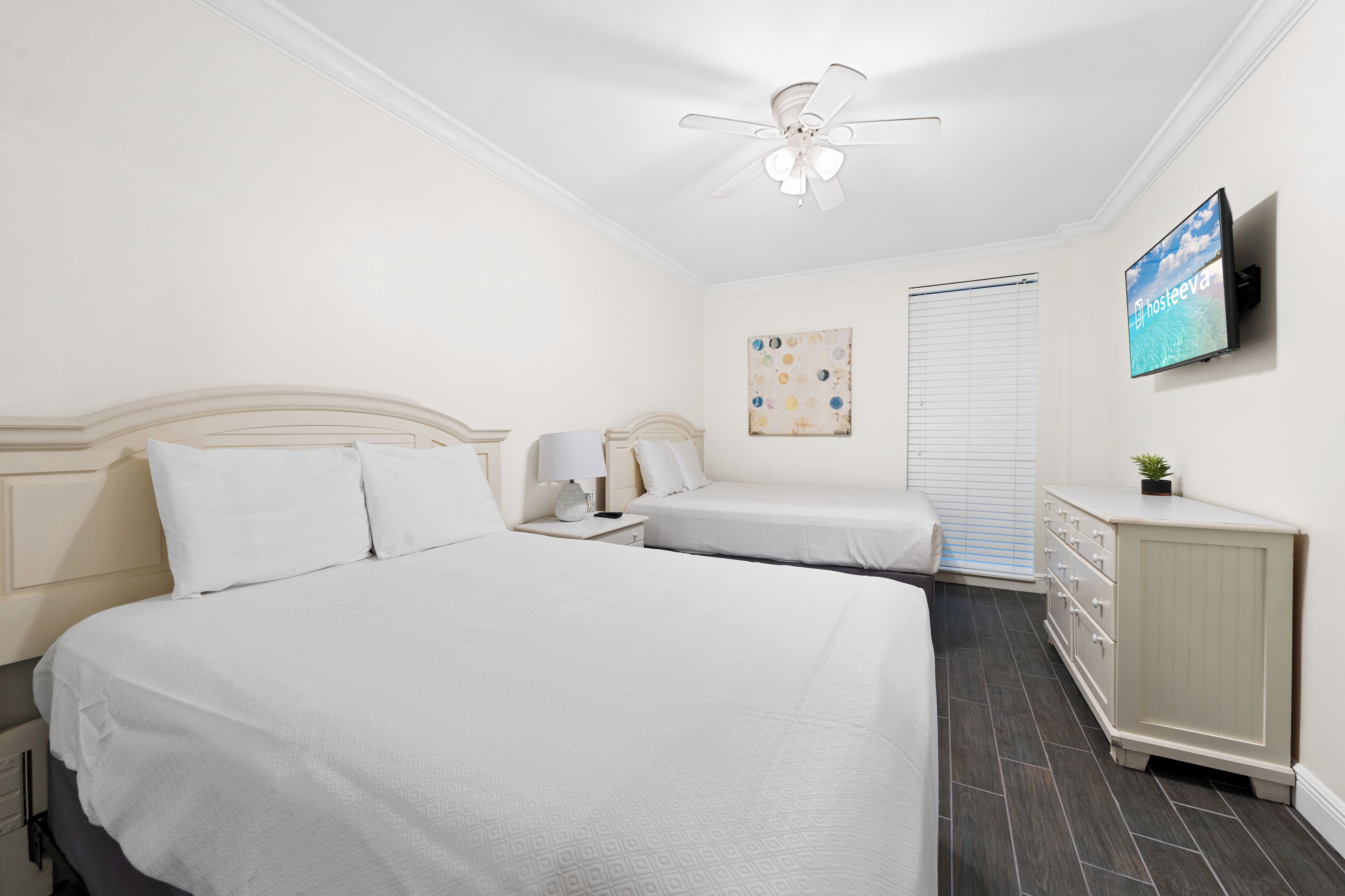 Habitación de hotel con dos camas, ventilador de techo, televisión, un cuadro y cómoda.