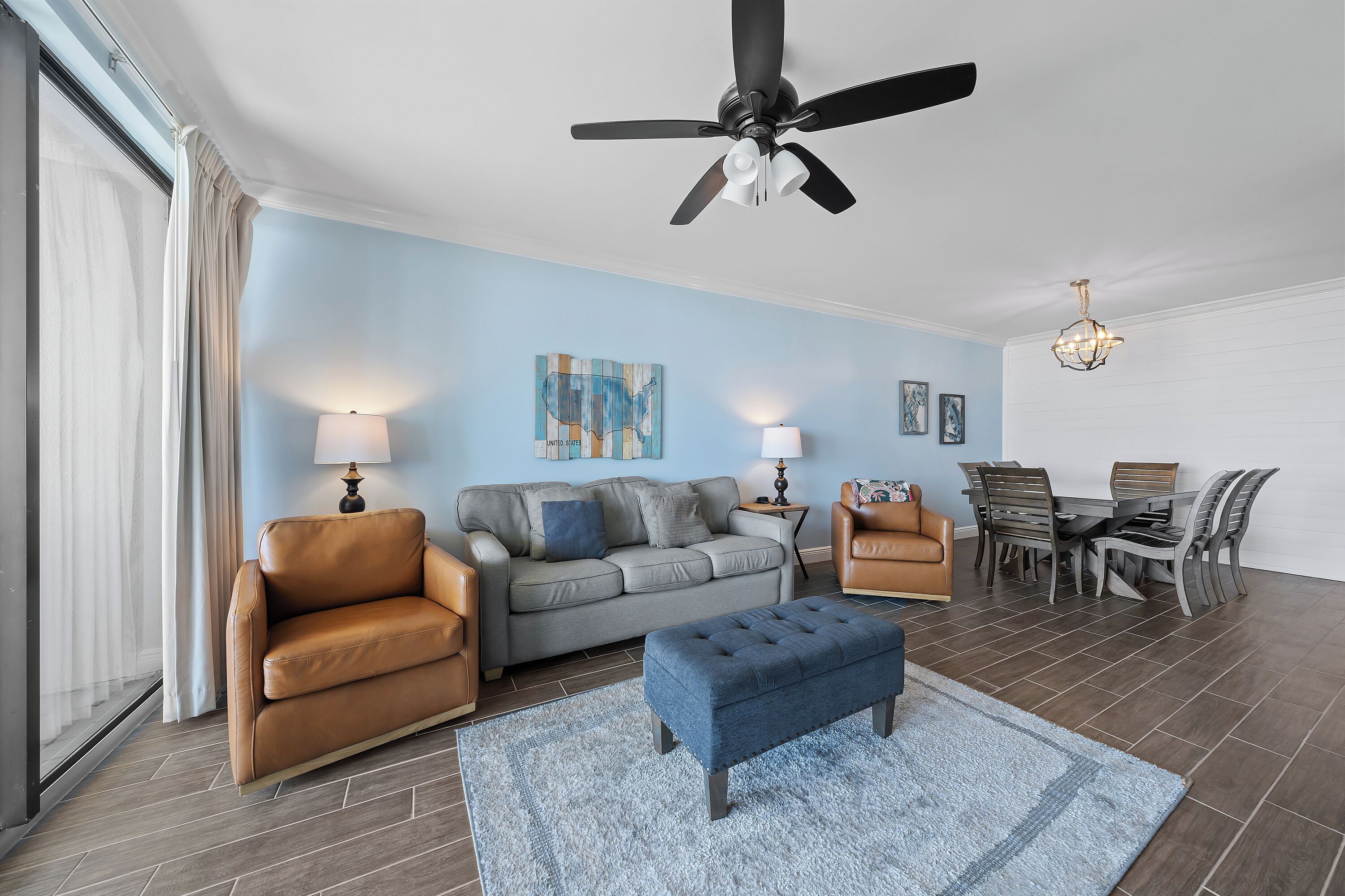 A living room with a grey sofa, two armchairs, a blue ottoman, a dining table with chairs, a ceiling fan, and two wall-mounted lamps.
