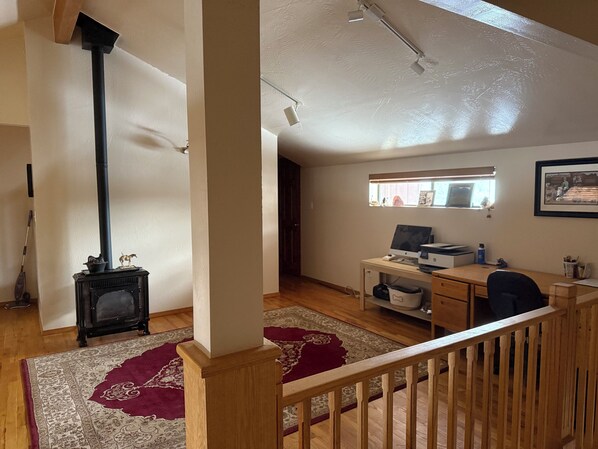 A room with a wooden desk, a computer, a window with blinds, a fireplace, and a red patterned rug.