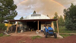 Eine überdachte Konstruktion mit einem blauen Traktor davor, umgeben von Bäumen und einem Regenbogen am Himmel.