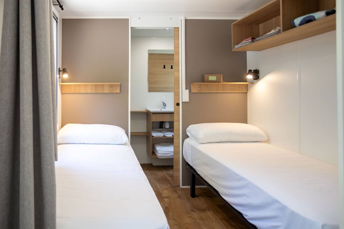 A hotel room with two single beds, a wooden wardrobe, a small bathroom, and a shelf with folded towels.