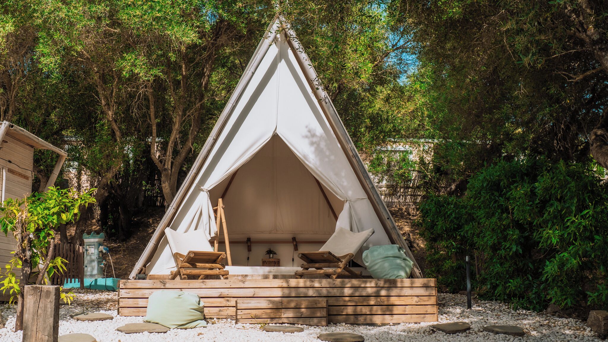 A triangular tent with a wooden platform and seating area, surrounded by trees and gravel.