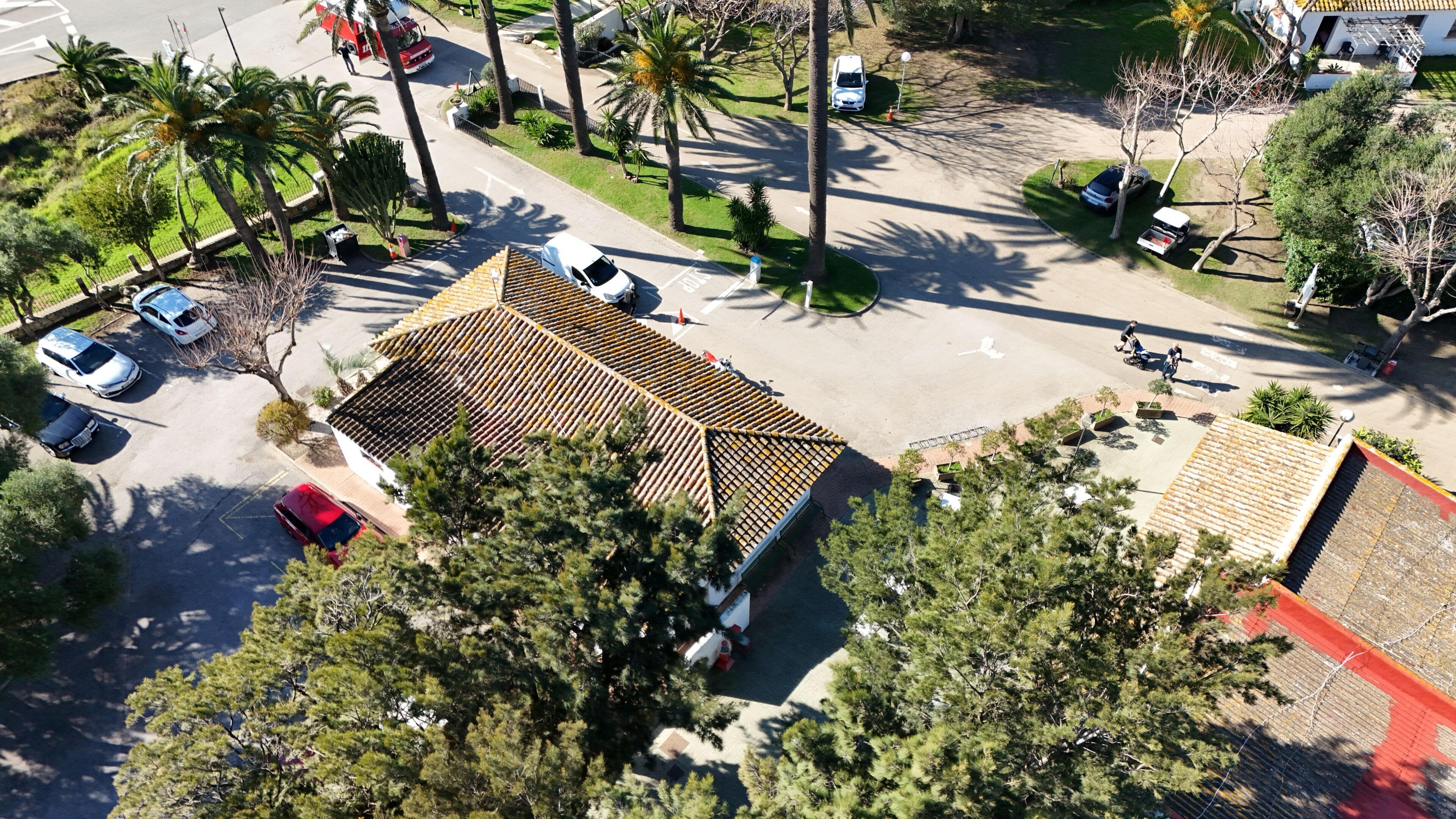 An aerial view of a parking lot with several cars, a building with a tiled roof, and surrounding greenery.
