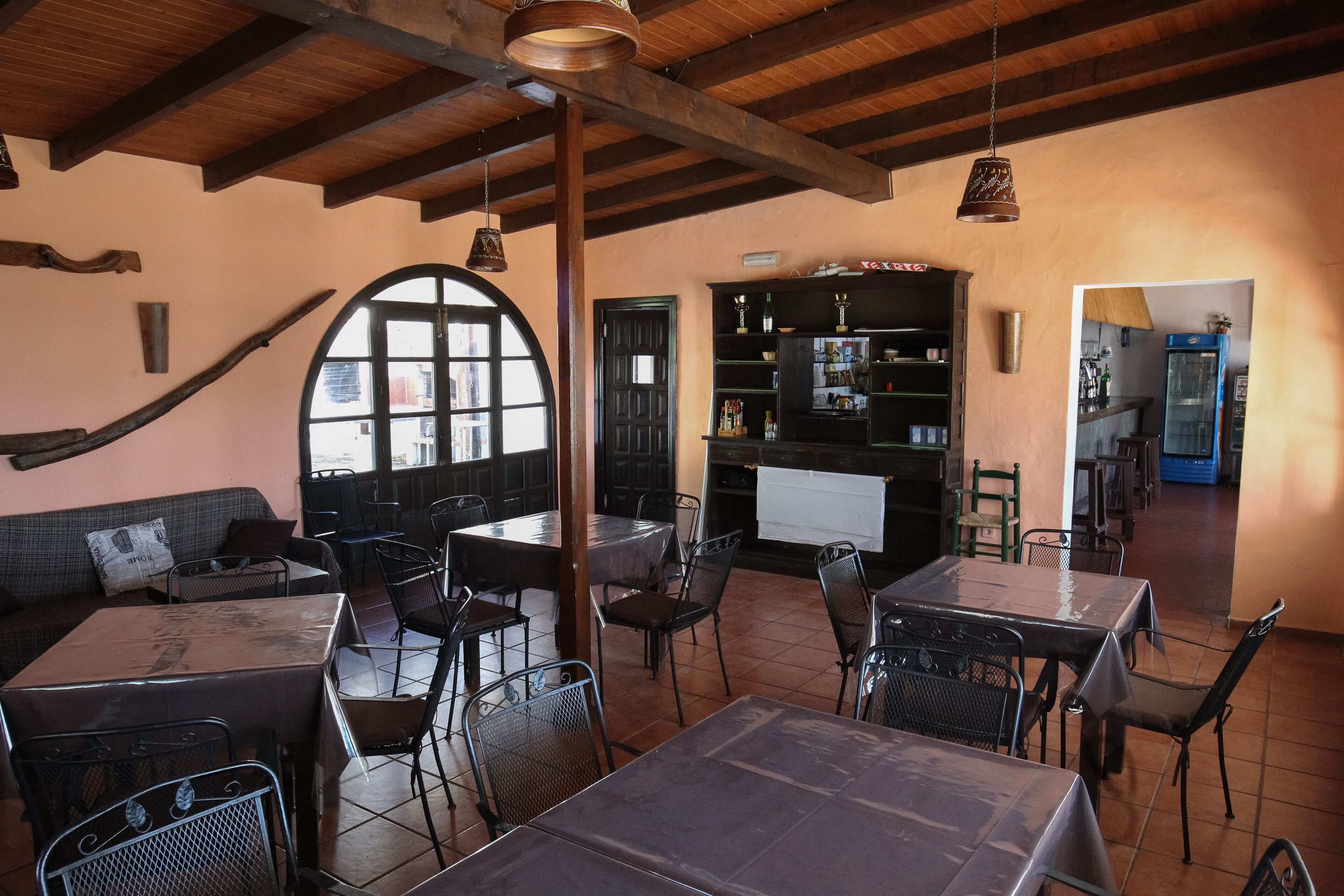 Un restaurante con vigas de madera, un gran ventanal arqueado y una estantería negra. Mesas y sillas dispuestas para comer.