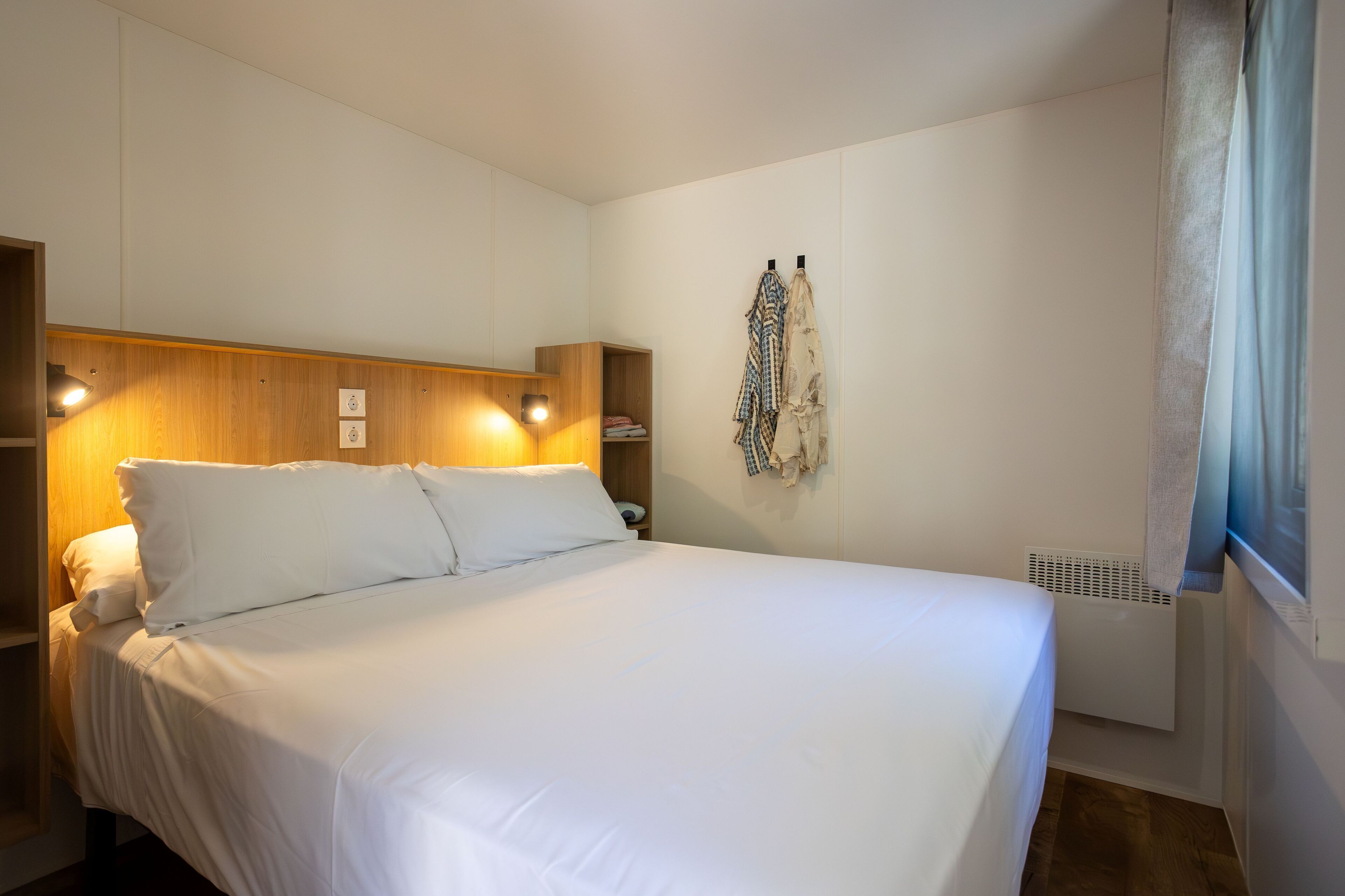 Una cama bien hecha con sábanas blancas en una habitación con cabecera de madera y estantes fijados a la pared.