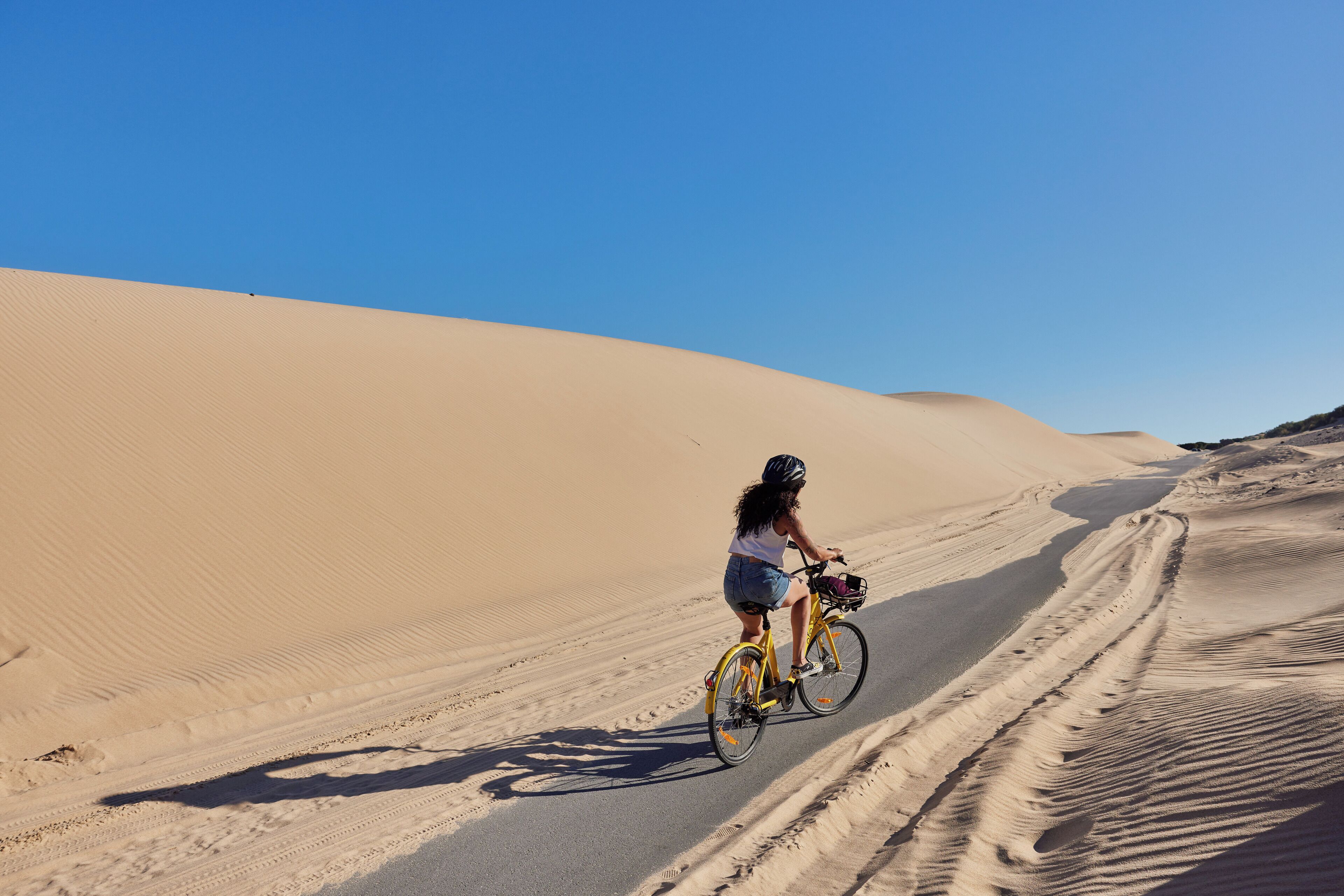 Una persona pedalea una bicicleta por un sendero arenoso, con dunas de arena al fondo.