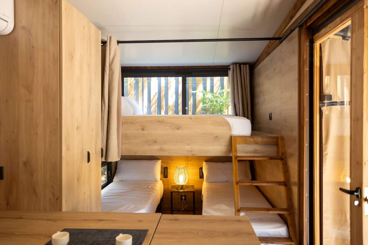 A compact, wooden-themed room with bunk beds, a small bedside table with a lamp, and a view through a window with curtains.