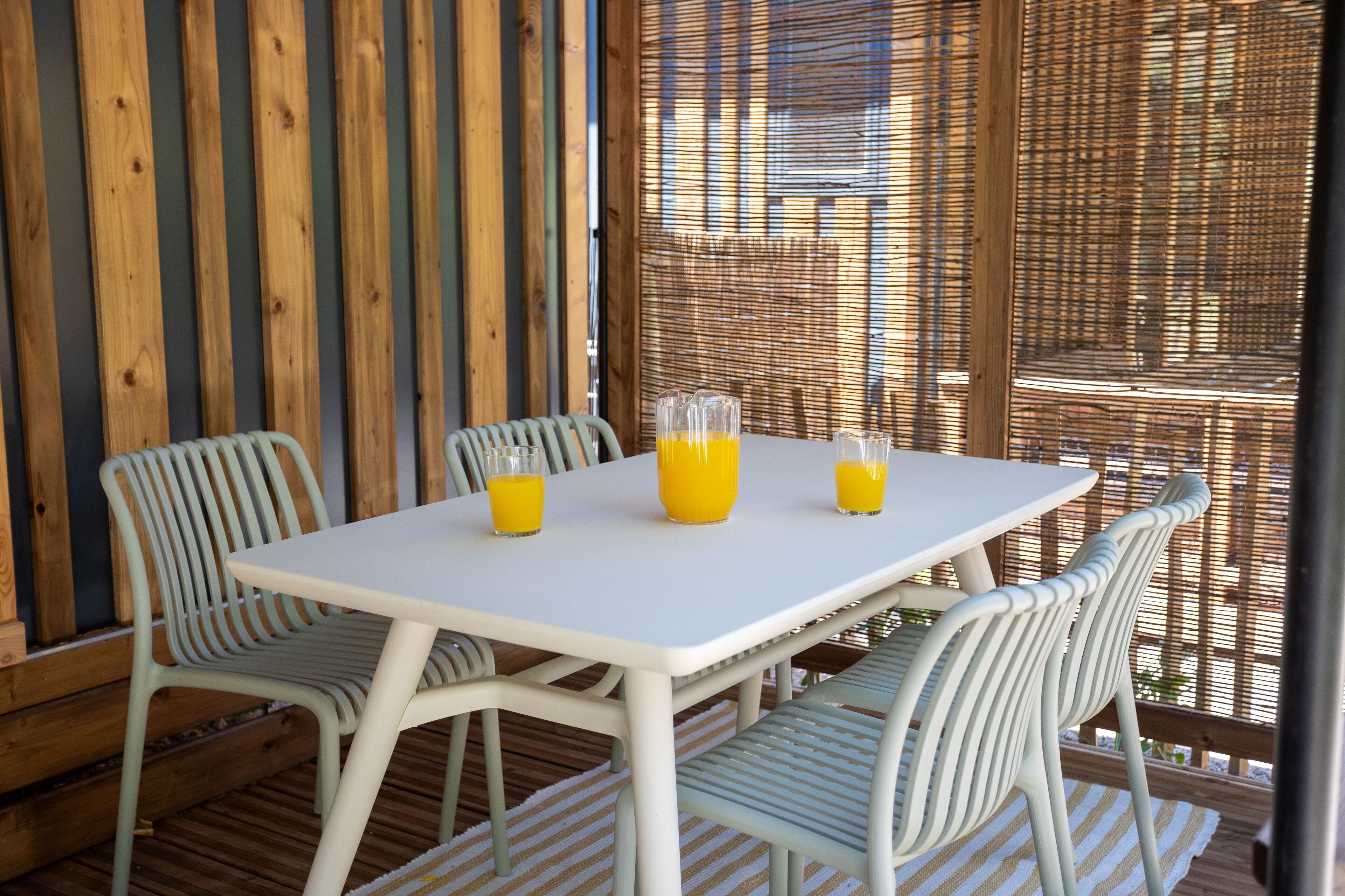 Uma mesa branca com quatro cadeiras, três copos de suco de laranja e um biombo de bambu ao fundo.
