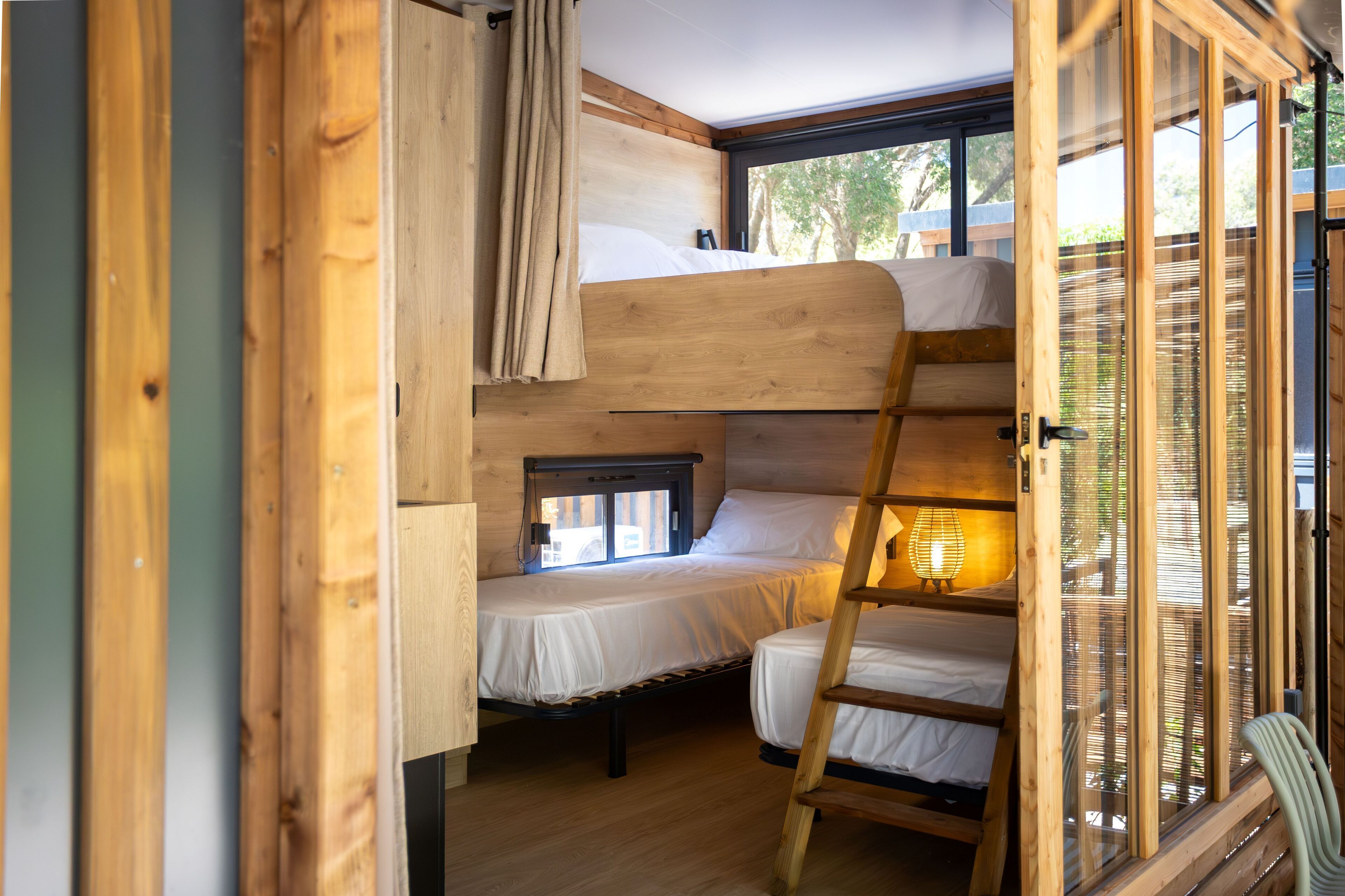 A compact, wooden interior with bunk beds and a ladder, featuring a window with blinds.