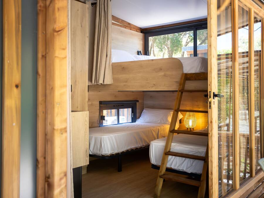 A compact, wooden interior with bunk beds and a ladder, featuring a window with blinds.