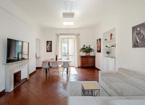 A living room with a sofa, a TV, a fireplace, a dining area with chairs, and a kitchen area with cabinets.