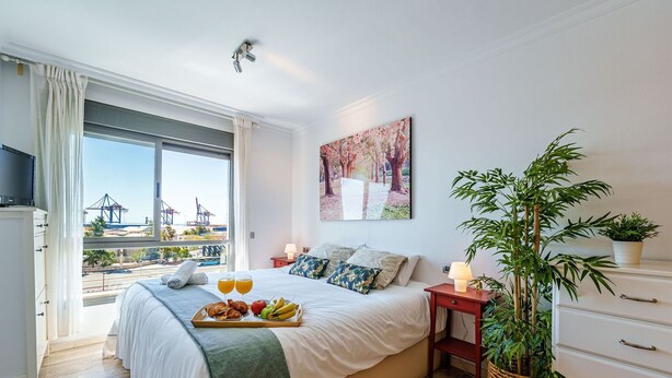 A hotel room with a large bed, a bedside table with a lamp, a window with a view of a port, and a painting on the wall.