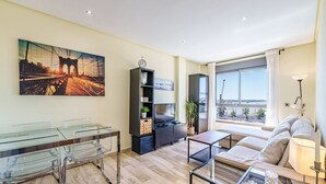 A modern living room with a glass dining table, a sofa, a TV, and a large window with a view of a bridge and construction.