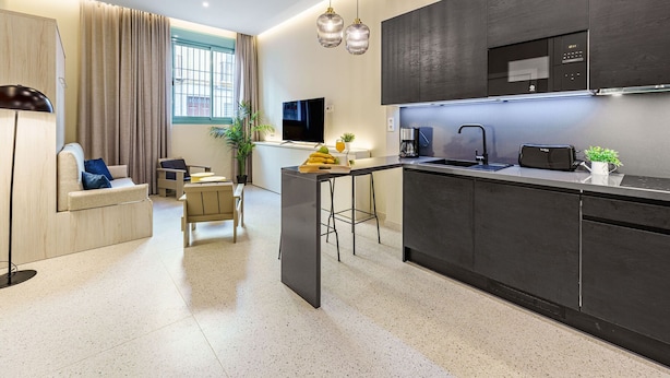Una cocina moderna con armarios oscuros, un microondas integrado y una cafetera. Hay una pequeña zona de estar con un sofá y una mesa de centro, y un amplio ventanal con cortinas.