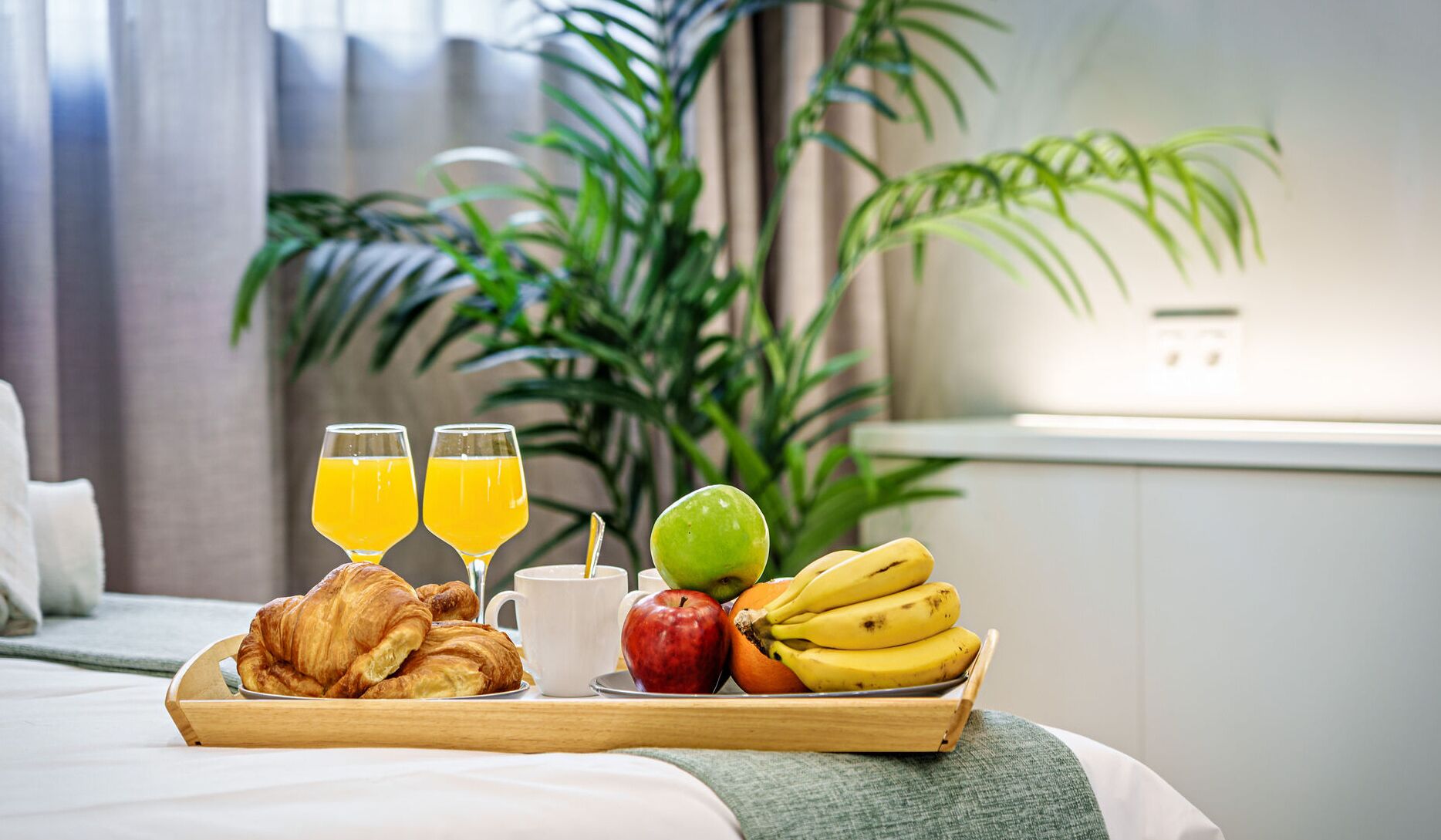 Ein Frühstücksbrett mit Croissants, Orangensaft, einer weißen Tasse, einem Apfel, einer Banane und einer grünen Frucht in einem Hotelzimmer mit einer Pflanze im Hintergrund.