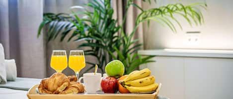 Un plateau de petit-déjeuner avec des croissants, du jus d’orange, une tasse blanche, une pomme, une banane et un fruit vert, posé sur un lit dans une chambre d’hôtel, avec une plante en arrière-plan.