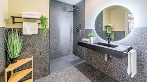 Un baño moderno con ducha de granito negro, espejo redondo y repisa de madera con toallas y plantas.