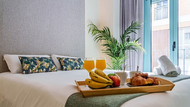 A neatly made bed with tropical patterned pillows, a tray with fruits, a plant, and a window with a view of a building. - Sunset Malaga Apartments - WIDE 1+2+3 (Málaga)