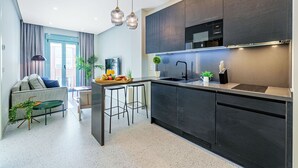 A modern kitchen with dark cabinetry, a stainless steel microwave, and a sink. There is a breakfast bar with stools and a small table with a tray of food and a plant. - Sunset Malaga Apartments - WIDE 1+2+3 (Málaga)