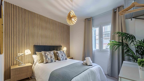 A modern bedroom with a wooden slat wall, a bed with white bedding and patterned pillows, bedside lamps, a bedside table, a large window with curtains, and a potted plant.