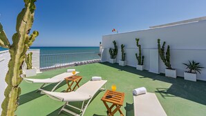 Terraza en la azotea con tumbonas, una mesa con una bebida y cactus en macetas frente a una pared blanca con vista al mar.