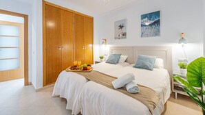 Habitación de hotel con un amplio armario de madera, cama con ropa blanca, dos almohadas y una bandeja con fruta y jugo. Hay luces fijadas en la pared y dos cuadros enmarcados.