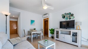 Una sala de estar moderna con un sofá, una mesa de centro de vidrio, un televisor sobre un mueble bajo y un ventilador de techo.