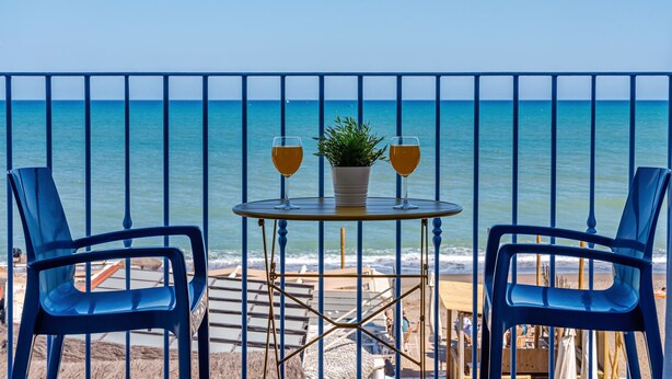 Ein Balkon mit zwei blauen Stühlen, einem kleinen runden Tisch und zwei Gläsern eines goldenen Getränks. Auf dem Tisch steht ein Blumentopf. Im Hintergrund Meer und ein sandiger Strand.
