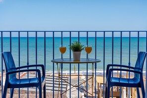 Un balcon avec deux chaises bleues, une petite table ronde et deux verres contenant une boisson dorée. Une plante en pot est posée sur la table. La vue embrasse l’océan et une plage de sable.