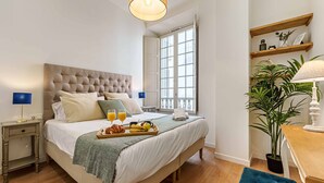 A hotel room with a large bed, a bedside table with a lamp, a tray with breakfast items, and a window with a view of buildings.