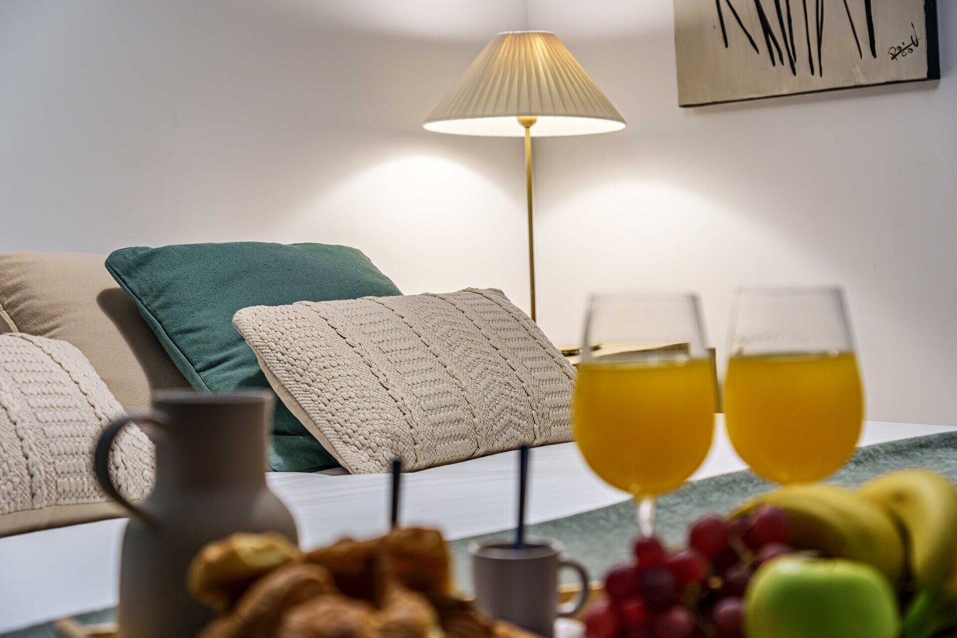 Habitació d'hotel ben ordenada amb un llit, almoinos, una làmpada, una testoseta, gots de suc i una safata de fruita.