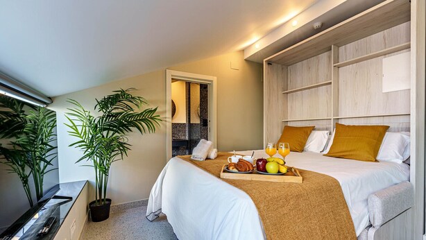 A hotel room with two beds, a TV, a potted plant, and a breakfast tray with fruits and pastries.