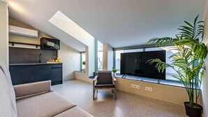 A modern apartment with a kitchenette, a dining area, a seating area with a sofa and a chair, a large window with a TV, and a potted plant.