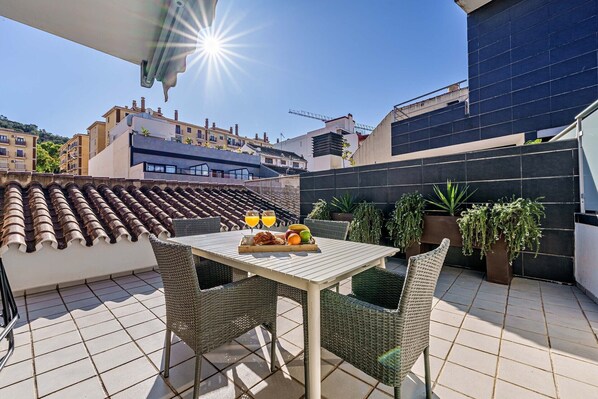 A rooftop terrace with a table set for two, surrounded by potted plants and a view of the cityscape.
