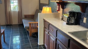 Uma cozinha compacta com piso de azulejos escuros, armários de madeira, micro-ondas, cafeteira e um pequeno espaço de estar com um banco e uma mesa.